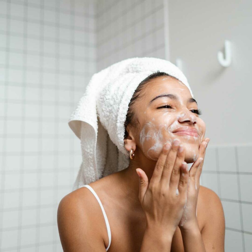 A joyful woman with a radiant smile on her face, lathering a facial cleanser onto her skin with her hands, as she enjoys her skincare routine.